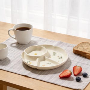 May include: A cream-colored divided plate with floral designs, a white mug, and a small cup sit on a striped placemat. Sliced strawberries, blueberries, and a piece of toast are also present. The plate has three sections for food.