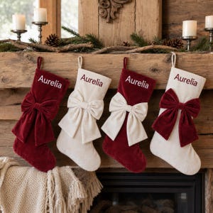 May include: Four Christmas stockings hanging in a row, alternating between deep red and cream velvet fabric. Each stocking features a large, matching velvet bow at the top and the name Aurelia embroidered in a contrasting color on the cuff.