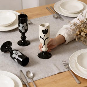 May include: Black and white ceramic vases with floral designs. One vase is held by a person. The vases are on a table set with plates, silverware, and a gray runner. The vases have a pedestal base.