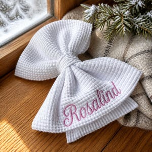 Peut inclure: Un nœud en tissu blanc à texture gaufrée portant le prénom Rosalind brodé en écriture cursive rose. Le nœud est composé d'un nœud central et de deux larges boucles repliées, présentant un motif en grille distinct sur toute la surface.