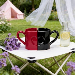 May include: A red and black ceramic mug set, designed to form a heart when joined. Each mug has a handle and a heart-shaped indentation. The mugs are on a small white table, with a bottle of water and flowers nearby.