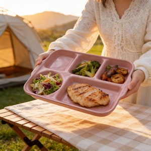 Puede incluir: Un plato rosa dividido con una pechuga de pollo a la parrilla, ensalada, brócoli y verduras asadas. El plato se sostiene al aire libre, con una tienda de campaña y luz solar en el fondo. El plato es rectangular con cinco compartimentos.