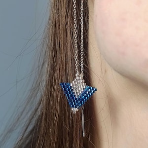Puede incluir: Pendientes colgantes con un diseño geométrico. Los pendientes tienen una cadena plateada y un colgante triangular hecho de cuentas azules y plateadas. Una pequeña barra plateada cuelga debajo. Los pendientes se muestran sobre un fondo borroso.