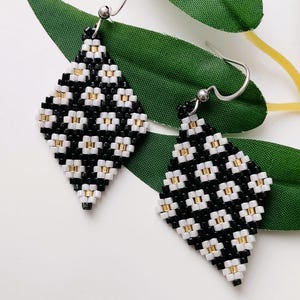 Puede incluir: Un par de pendientes de diamantes con cuentas. Los pendientes presentan un patrón de cuentas negras y blancas con detalles dorados. Tienen ganchos plateados y se muestran sobre un fondo blanco con hojas verdes.