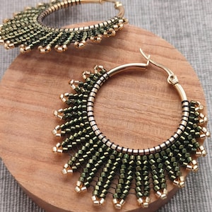 Puede incluir: Pendientes de aro dorados con un diseño de cuentas en tonos verdes y dorados. Los pendientes presentan un patrón similar a una franja de pequeñas cuentas, creando un aspecto texturizado y decorativo. Los aros se muestran sobre una superficie de madera.