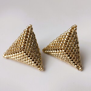 以下が含まれることがあります： 金色の三角形のスタッドピアス。各イヤリングは、小さく均等に配置されたビーズで構成されており、テクスチャード加工された表面を作り出しています。イヤリングは無地の白い背景に配置されています。