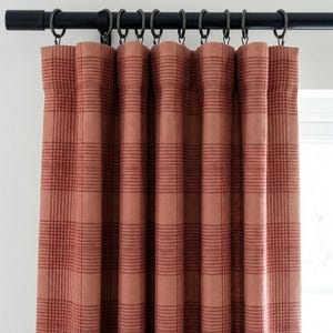 May include: A set of terracotta-colored plaid curtains hanging from a black metal rod with matching rings. The curtains are made of a textured fabric and are partially covering a window.
