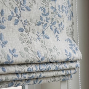 Puede incluir: Un estor plegable de tela beige claro con un patrón bordado de ramas y hojas delicadas en tonos azul y verde salvia. El cubrimiento de ventana presenta un diseño plegado con una cadena de cuentas blanca en el lateral para su manejo.