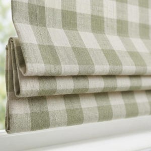 May include: Close-up of a green and white checkered Roman shade. The fabric is folded in horizontal layers, revealing the pattern. The shade is mounted on a white window frame, with a glimpse of the outdoors visible in the background.