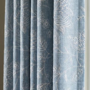 May include: A light blue curtain panel featuring a white embroidered floral and botanical pattern. The fabric has a visible weave texture and hangs in vertical folds, displaying a repeating design of leaves, stems, and blossoms throughout.