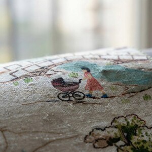 May include: Close-up of a fabric with a printed scene. A woman in a red dress pushes a brown stroller along a path. The scene includes a bridge, trees, and a light blue area, all in muted colors.