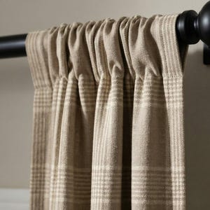 May include: Close-up of a beige plaid curtain panel hanging from a black rod. The curtain has a textured appearance with a repeating pattern of light beige and off-white stripes. The rod has a decorative finial.
