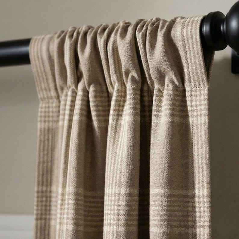 May include: Close-up of a beige plaid curtain panel hanging from a black rod. The curtain has a textured appearance with a repeating pattern of light beige and off-white stripes. The rod has a decorative finial.
