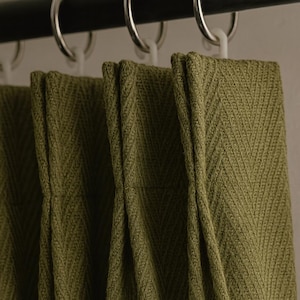 Pode incluir: Close-up de cortinas verde-oliva com um padrão de espinha de peixe texturizado. O tecido é drapeado e pendurado em uma barra preta com anéis prateados e ganchos de plástico brancos. As cortinas são feitas de um material espesso e tecido.