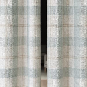 May include: Close-up of two light-colored plaid curtains. The curtains feature a pattern of light blue, beige, and white squares. The fabric has a textured appearance. The curtains are hanging, with a dark space between them.