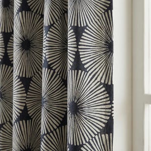 Pode incluir: Cortinas estampadas em azul marinho e creme. O tecido apresenta um design repetitivo de raios de sol estilizados ou padrões radiais. As cortinas estão penduradas em dobras verticais, com um vislumbre de uma janela ao fundo.