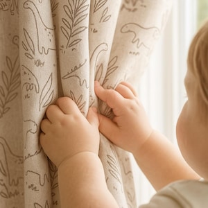 Puede incluir: Cortina beige con un patrón repetido de dinosaurios y follaje en marrón. Las manos de un niño sostienen la cortina. La tela tiene un aspecto suave y texturizado. Ideal para la habitación de un niño.