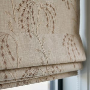 May include: A beige roman shade featuring an embroidered pattern of delicate, drooping floral stems in a darker brown thread. The fabric has a visible woven texture, and the shade is constructed with horizontal folds that create a layered appearance.