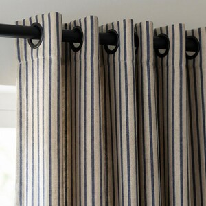 Op de afbeelding: Close-up van gestreepte gordijnen. De gordijnen zijn beige met verticale marineblauwe strepen. Ze hangen aan een zwarte roede met zwarte ogen. De stof heeft een getextureerd uiterlijk.