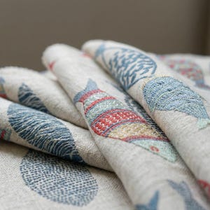 May include: Close-up of folded fabric with embroidered fish designs. The fish are in shades of blue, red, and green, set against a neutral background. The fabric appears to be linen or a similar woven material, with a textured surface.