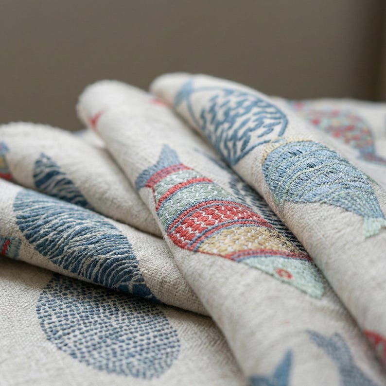 May include: Close-up of folded fabric with embroidered fish designs. The fish are in shades of blue, red, and green, set against a neutral background. The fabric appears to be linen or a similar woven material, with a textured surface.