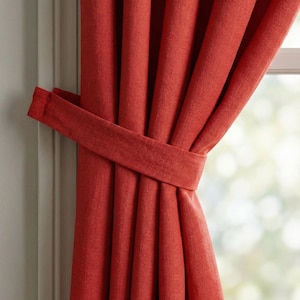 Puede incluir: Primer plano de cortinas rojas con una cinta a juego. Las cortinas están hechas de una tela texturizada y están parcialmente corridas, revelando un vistazo de una ventana y luz natural. La cinta está envuelta alrededor de las cortinas.