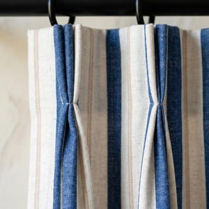 Puede incluir: Primer plano de un par de cortinas a rayas. Las cortinas presentan rayas verticales en blanco, beige y azul. La tela está fruncida en la parte superior y cuelga de aros metálicos negros.