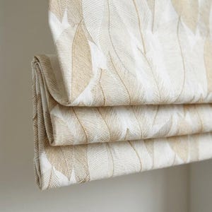 May include: Close-up of a folded Roman shade with a repeating feather pattern. The fabric features a cream-colored background with large, light brown feather designs. The shade is folded in horizontal layers, showcasing the pattern.