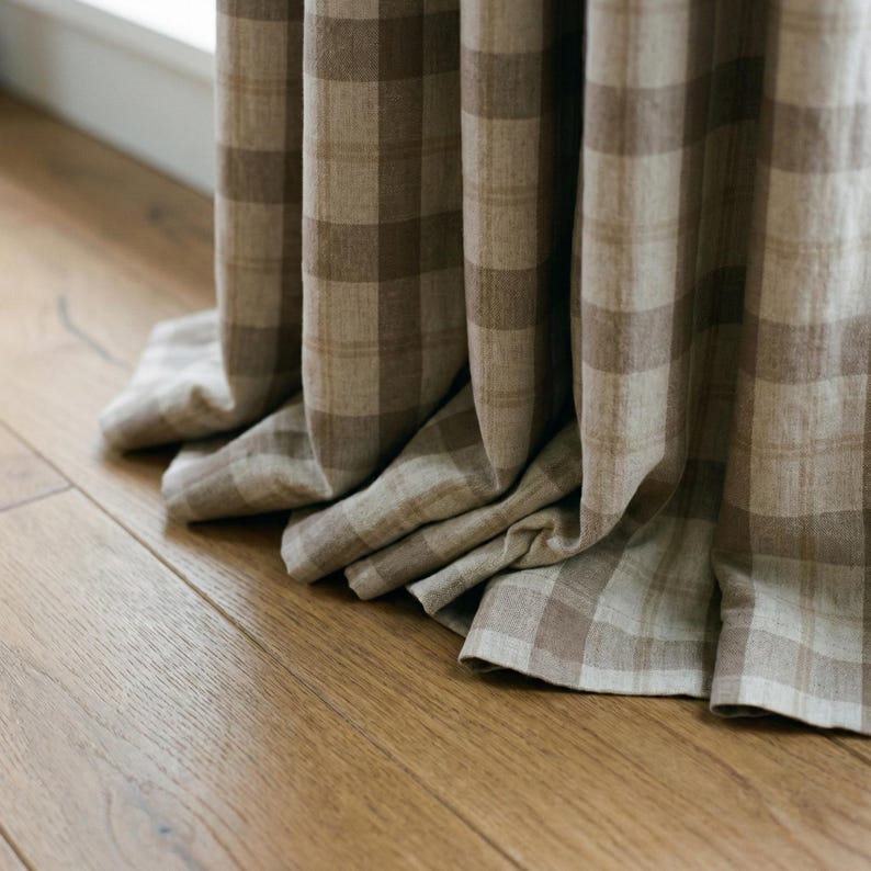 May include: Close-up of beige and brown plaid curtains. The curtains are made of a soft fabric and are draped in folds. The curtains are hanging in front of a window, and the floor is made of wood.