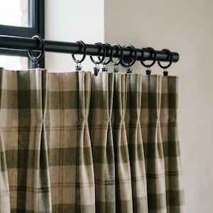 May include: Green and tan plaid curtains hanging from a black metal rod with black rings. The curtains are pleated and hang in vertical folds. The window is visible in the background.