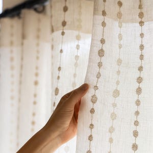 Könnte beinhalten: Beige Leinen-Vorhänge mit einem sich wiederholenden Muster aus gestickten Kreisen in einem dunkleren Beigeton. Die Vorhänge sind halbtransparent und lassen Licht durchscheinen. Eine Hand hält sanft den Rand des Vorhangs.