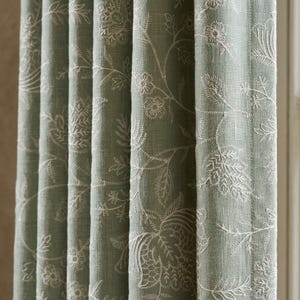 May include: A sage green curtain panel featuring a white embroidered botanical pattern of flowers and leaves. The fabric has a visible weave texture and hangs in vertical folds, displaying a repeating floral design across the entire surface.