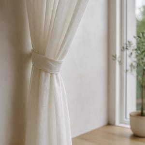 Puede incluir: Cortina de lino color crema atada con una cinta de tela a juego. La cortina cuelga frente a una pared de color claro, con una vista de una ventana y una planta en maceta en el fondo. Se aprecia la textura y los pliegues de la cortina.