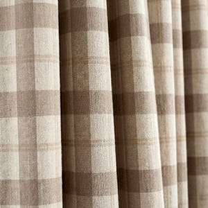 May include: Close-up of beige and brown plaid fabric curtains. The curtains have a large, square check pattern in varying shades of beige and brown. The fabric appears to be a woven material, with visible texture and slight folds. The curtains are hanging vertically.