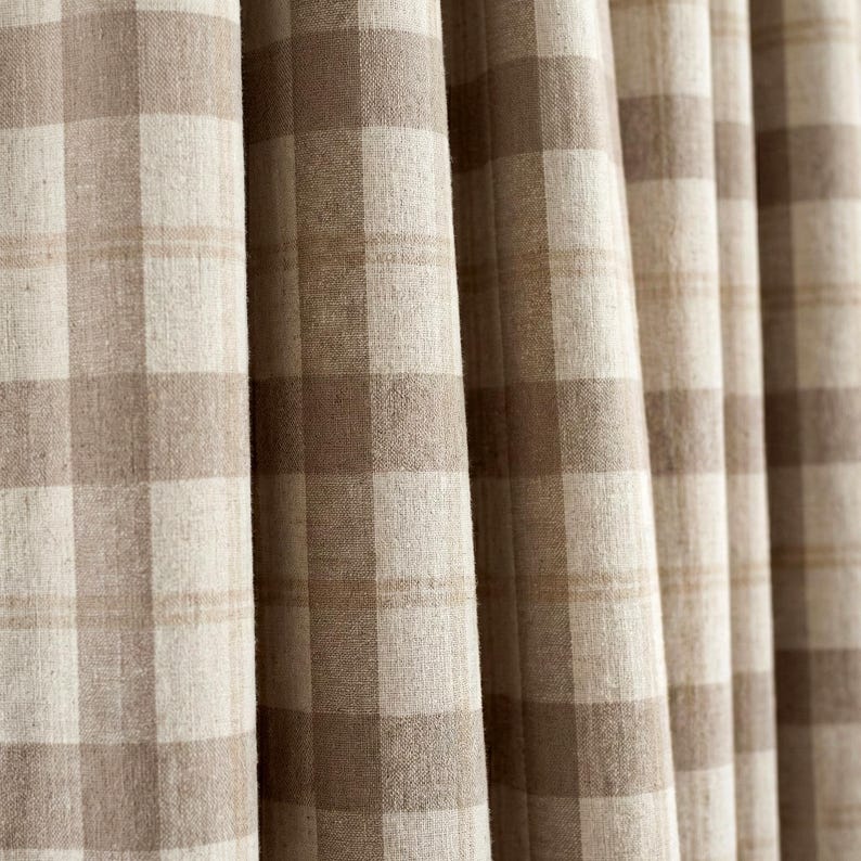 May include: Close-up of beige and brown plaid fabric curtains. The curtains have a large, square check pattern in varying shades of beige and brown. The fabric appears to be a woven material, with visible texture and slight folds. The curtains are hanging vertically.