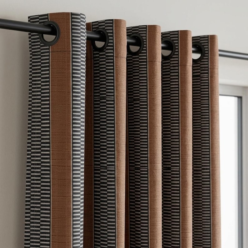Pu&ograve; includere: Un set di tende in tessuto testurizzato color marrone ruggine con pannelli verticali a righe geometriche bianche e nere. Le tende sono appese a un bastone in metallo nero tramite occhielli circolari integrati sul bordo superiore.