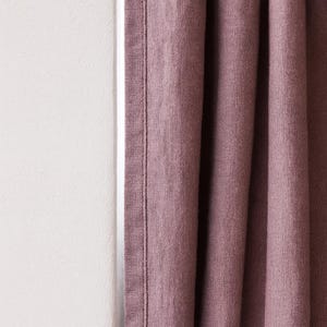 May include: Close-up of a dusty rose-colored curtain panel with vertical folds. The fabric has a textured appearance and is sewn with a vertical seam. A white wall is visible to the left of the curtain.