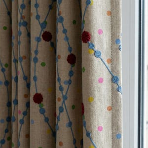 Könnte beinhalten: Beige Leinen-Vorhänge mit einem sich wiederholenden Muster aus blauen gestickten Linien und bunten Tupfen. Das Design umfasst rote, blütenartige Akzente. Die Vorhänge hängen vor einem Fenster.