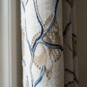 Puede incluir: Primer plano de una cortina de tela beige con un diseño de conchas marinas. El diseño presenta conchas bordadas en tonos marrones y beige, con detalles bordados en azul y azul claro. La cortina está hecha de una tela texturizada.
