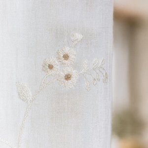 Könnte beinhalten: Nahaufnahme eines weißen Stoffes mit gesticktem Blumendesign. Die Stickerei zeigt drei gänseblümchenartige Blüten mit beigefarbenen Zentren und zarten Blättern. Der Stoff hat ein strukturiertes Aussehen.