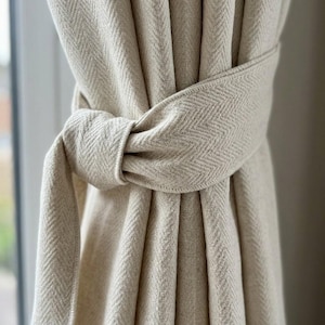 May include: Close-up of beige herringbone patterned curtains tied with a matching fabric belt. The curtains are made of a thick, textured material and are partially blocking a window. The tie is knotted in the centre.