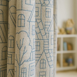 May include: Beige fabric curtain with a repeating pattern of blue embroidered houses and trees. The curtain has a textured appearance and is partially folded, revealing the design's detail. The design includes arched windows and pitched roofs.