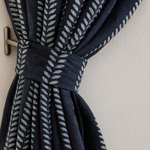 Op de afbeelding: Marineblau gordijn met een herhalend patroon van lichtgrijze bladvormen. Het gordijn wordt vastgehouden met een bijpassende stoffen band, en een metalen haak is zichtbaar aan de muur. De stof heeft een zachte, vloeiende uitstraling.