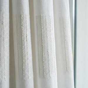 May include: Close-up of white linen curtains with vertical textured stripes. The curtains are made of a light-coloured fabric and hang in front of a window. The textured stripes add a subtle pattern to the otherwise plain design.