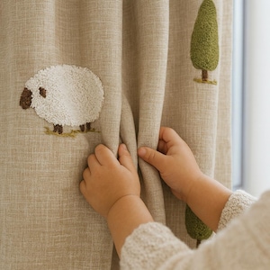 May include: Beige linen curtains with embroidered sheep and tree designs. The sheep has a white body, brown face, and is standing on green grass. A green tree is also embroidered on the curtain. The curtains are being held open by a person.