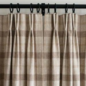 May include: Close-up of beige and brown plaid curtains hanging from a black metal rod with black rings. The curtains are pleated and have a classic, neutral design. The fabric appears to be a woven material.