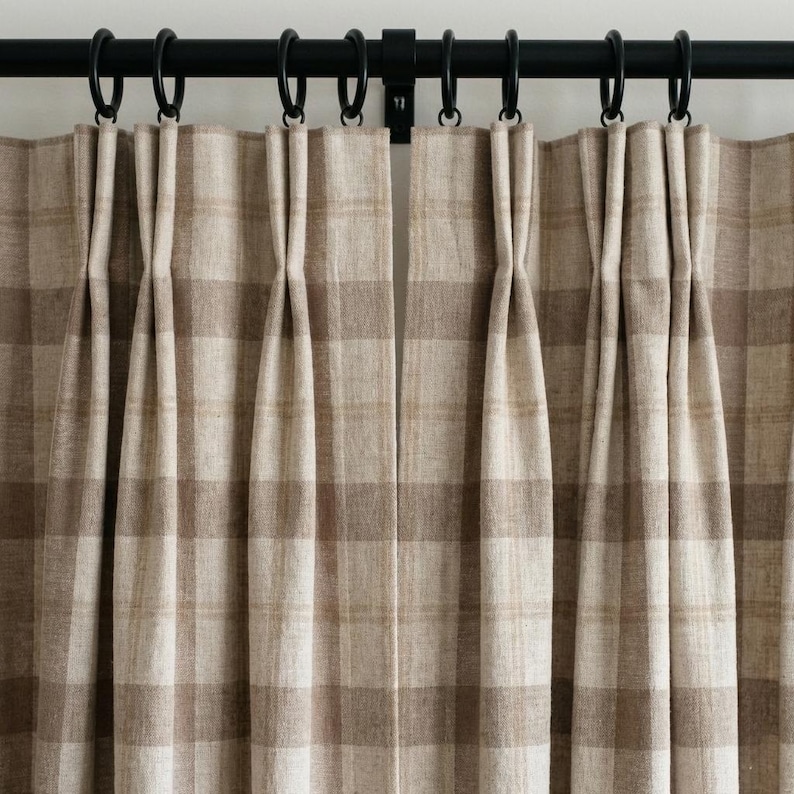 May include: Close-up of beige and brown plaid curtains hanging from a black metal rod with black rings. The curtains are pleated and have a classic, neutral design. The fabric appears to be a woven material.