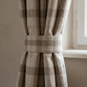 May include: Beige and brown plaid curtains held by a matching fabric tie-back. The curtains are made of a textured material and hang in soft folds. A white window frame is visible in the background, providing natural light.