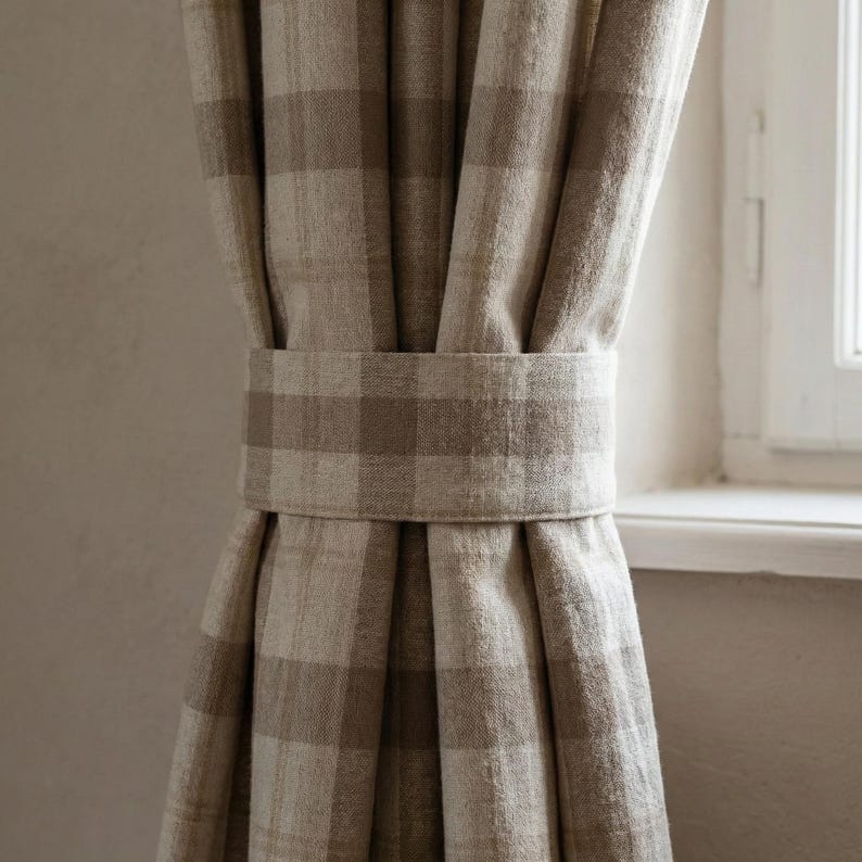 May include: Beige and brown plaid curtains held by a matching fabric tie-back. The curtains are made of a textured material and hang in soft folds. A white window frame is visible in the background, providing natural light.
