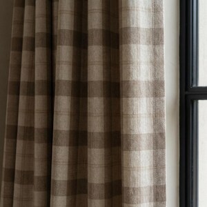 May include: Close-up of a brown and beige plaid curtain panel. The fabric has a textured appearance and is hanging near a window with a black frame. The curtain is made of a woven material.